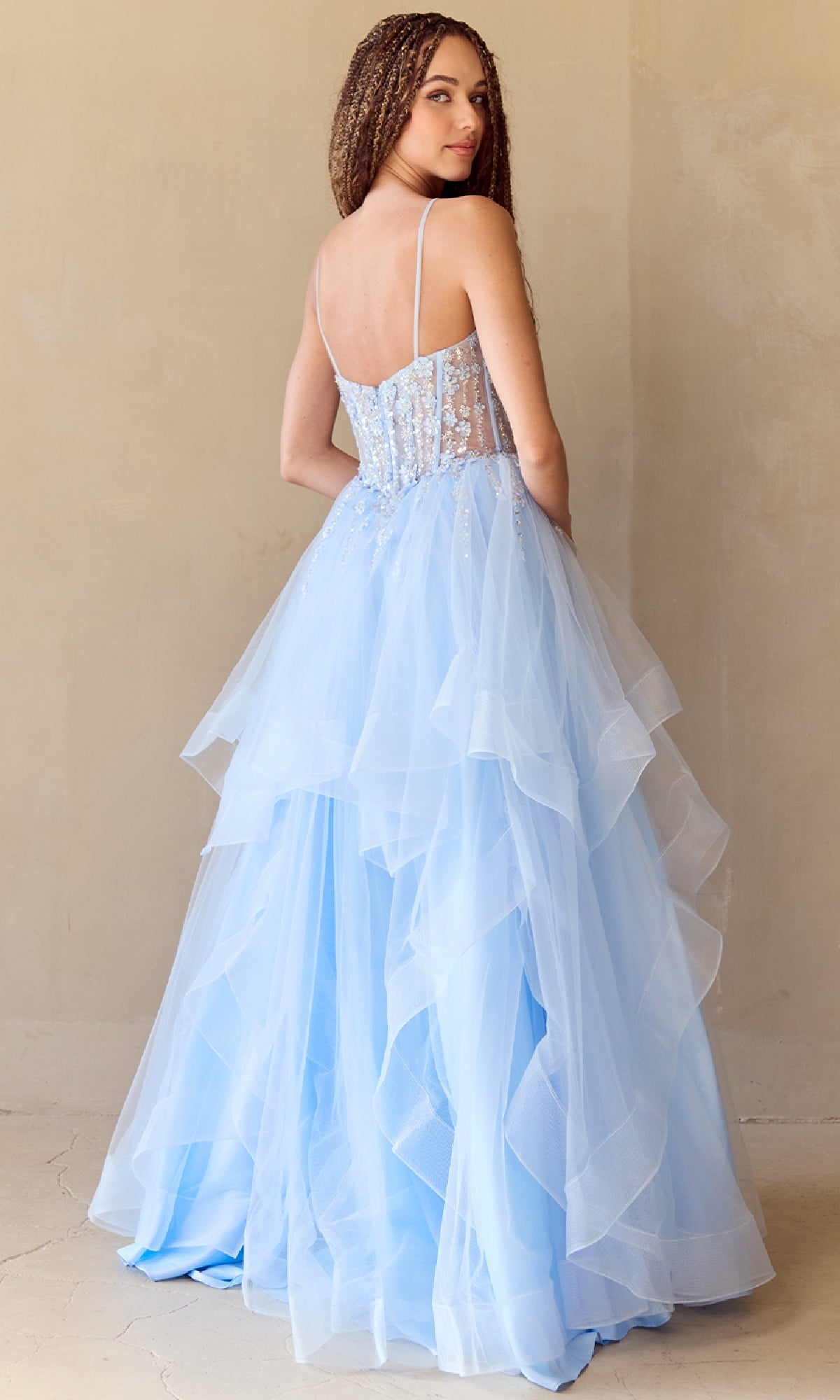 A woman stands indoors facing away from the camera, wearing the Amelia Couture 5068 Long Glitter Prom Ball Gown—a light blue, layered tulle dress with thin straps and a semi-sheer embellished bodice.